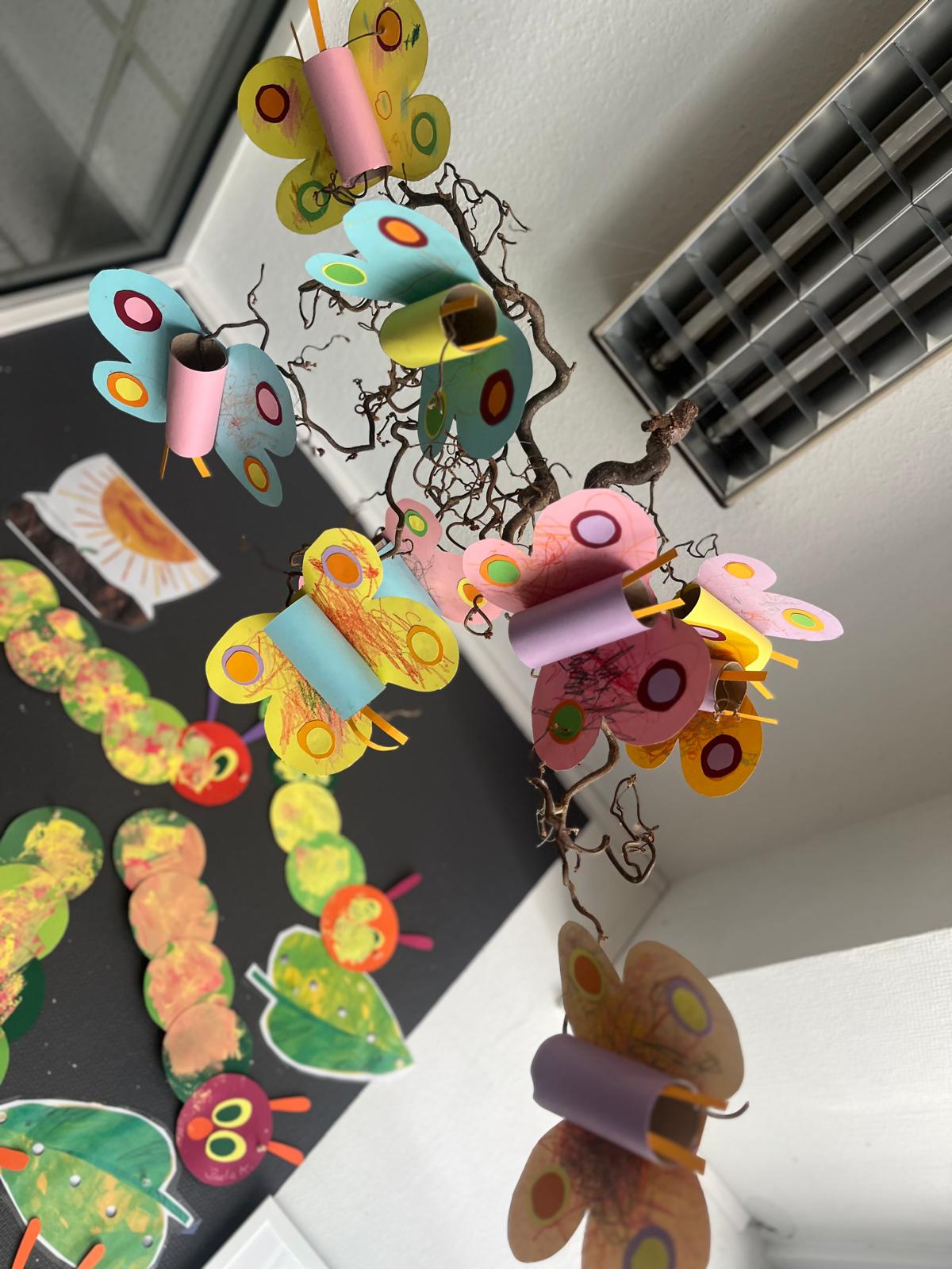 Bunte Schmetterlinge aus Papierrollen hängen an einem Zweig, dahinter Raupen an der Wand