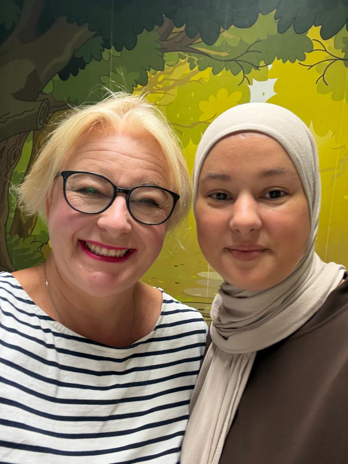 Daniela Brekalo und Najat Zarouali lächeln gemeinsam in die Kamera, hinter ihnen ein gemalter Wald